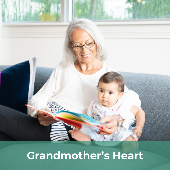 A senior women sits on a couch reading to a baby in her lap.There is a text bar on the bottom of the image that reads Grandmother's Heart.